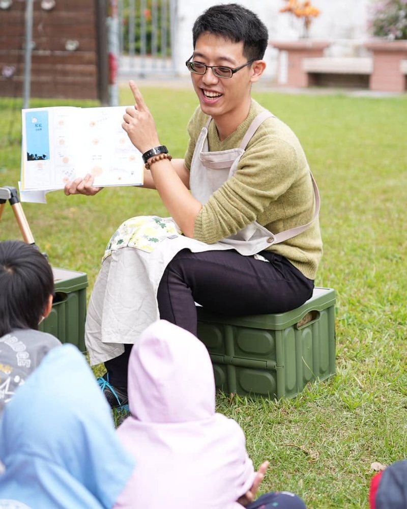 Awei sitting on the grass reading a picture book to children