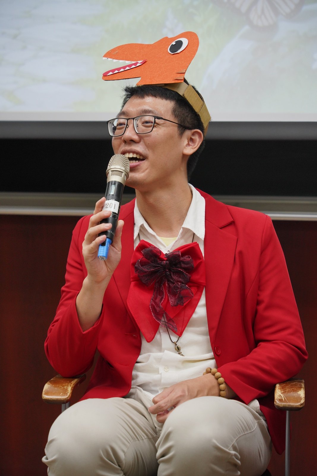 講師穿著紅色西裝、戴著恐龍頭飾，拿著麥克風演出故事