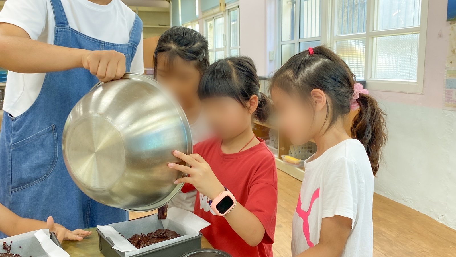 穿圍裙的大人傾斜鋼盆倒麵糊,孩子扶著盆邊看麵糊流進烤模